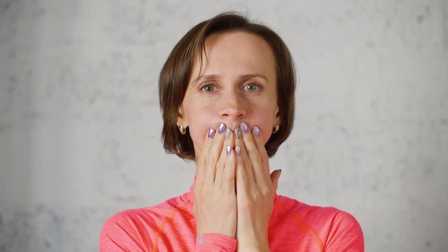 Woman does facial gymnastics exercises, puffs up her cheeks and clamps her lips with hands. Portrait of young woman covering her mouth with her hands. Wellness facial yoga against wrinkles.
