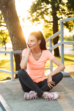Image of young fitness girl in sportswear looking aside while sitting at sports ground