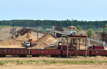 Fototapeta premium View on the mining quarry. Plant for the production sand and gravel for the construction industry. Mechanical machine, conveyor belt for crushing stone. Dump trucks transporting sand in the open-pit.