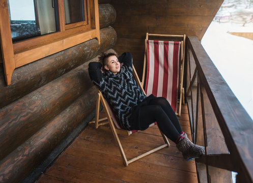 Happy woman rests in chaise lounge in the winter wooden chalet dressed in knitted sweater. Winter vacation