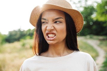 Photo of displeased woman wearing straw hat showing her teeth while walking in green park