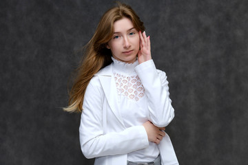 Portrait to the waist of a young pretty brunette girl woman with beautiful long hair on a gray background in a white jacket. He talks, smiles, shows his hands with emotions in various poses.