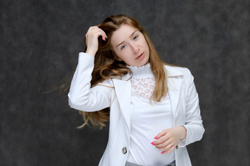 Portrait to the waist of a young pretty brunette girl woman with beautiful long hair on a gray background in a white jacket. He talks, smiles, shows his hands with emotions in various poses.