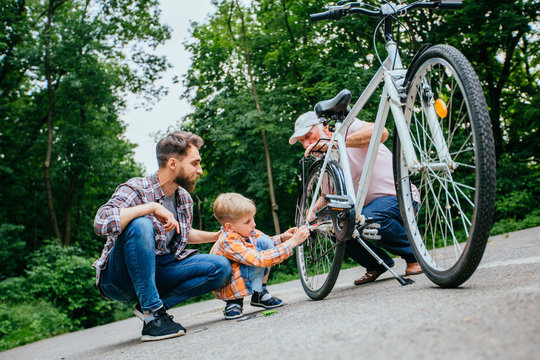 Father Teaching Son Reparing Bike Outdoor. Dad, Grandad And School Age Grandson Son Fixing Bicycle At Summer Green Park Outside. Happy Family Relation Different Generation Concept.