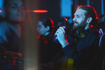 Smiling satisfied young bearded gamer with headphones on neck sitting in comfortable armchair and drinking energy beverage while playing video game