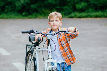 Child boy in orange shirt carries a big bike in the summer park outdoor.