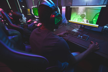 Rear view of addicted gamers sitting at table and looking at monitors while playing online multiplayer game