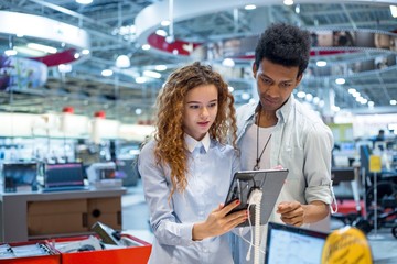 Red-haired girl with glasses with an African guy in an electronics store standing in front of the stand choosing a tablet computer to buy
