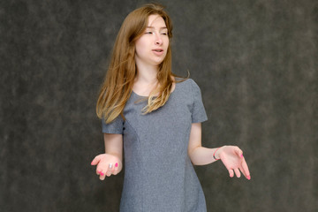 Portrait to the waist of a young pretty brunette girl woman with beautiful long hair on a gray background in a gray dress. He talks, smiles, shows his hands with emotions in various poses.