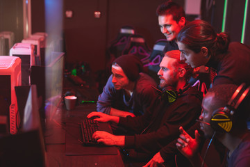 Group of focused young men standing together in front of computer monitor and playing video game, friends assisting guy to pass network game
