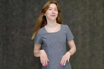 Portrait to the waist of a young pretty brunette girl woman with beautiful long hair on a gray background in a gray dress. He talks, smiles, shows his hands with emotions in various poses.