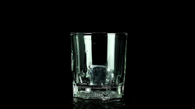 Filling ice cubes and whiskey in empty glass on dark background. Whisky drink at bar counter.