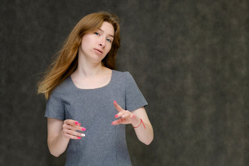 Naklejka premium Portrait to the waist of a young pretty brunette girl woman with beautiful long hair on a gray background in a gray dress. He talks, smiles, shows his hands with emotions in various poses.