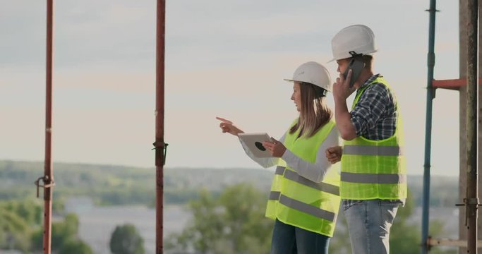Engineers designers stand on the roof of the building under construction and discuss the plan and the progress of construction using a tablet and mobile phone