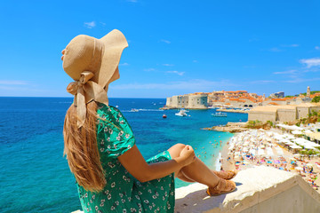Beautiful young woman with hat sitting on wall looking at stunning panoramic village of Dubrovnik in Croatia, Europe
