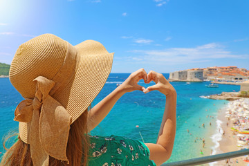 Young beautiful woman in Dubrovnik making hands shaped heart on spring summer warm sunny day. Girl with hat happy outdoors in her travel in Southern Croatia, Europe. - Powered by Adobe