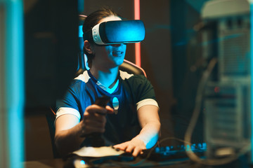 Fototapeta na wymiar Smiling young guy in virtual reality goggles sitting at table and using joystick while having 3D experience