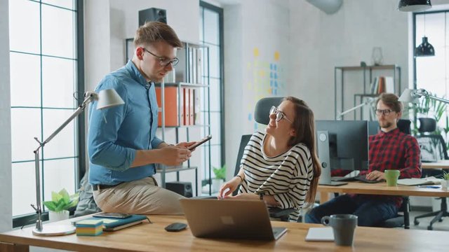 Male Colleague Sits on a Table in Front of a Young Female Coworker while Holding a Tablet Computer with Business Notes