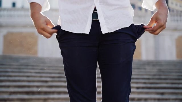 Sad man staying on the stairs outdoors and touching his trousers pockets. Young disappointed tourist turning out his empty pockets on the street. No money inside the pockets. Close-up shot of looking