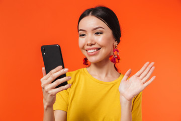 Happy young woman posing isolated over orange wall background talking by mobile phone take a selfie waving.