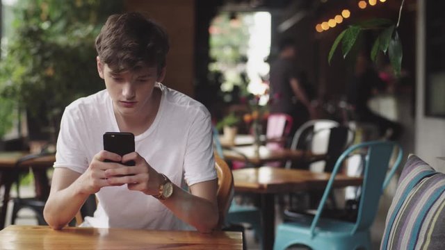 Young Man surfing internet in mobile phone, handy touch and sending text message in cafe. Handsome Teenager chatting in mobile app in public place. 
