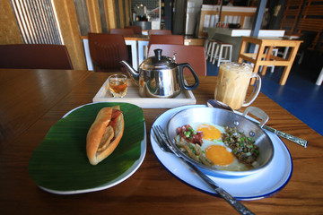 Indochina pan-fried egg with toppings with Baguette bread sandwich with cheese, ham on fresh Green banana leaf and ice coffee, tea hot on wooden table in homemade Thai style