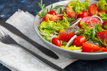 Salad of tomato, onion, pepper, arugula and chia seeds. Dressed with soy sauce and olive oil. Near the stick with dressing. On a dark background. Metal fork and knife. 