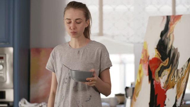 Young female painter in stained t-shirt eating cereals from bowl and using laptop while standing in home art studio