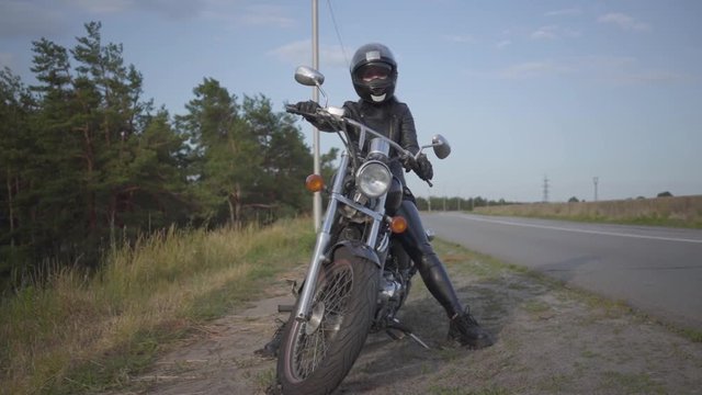 Front view of the successful confident young woman in leather dress sitting on her bike at the road. Hobby, traveling and active lifestyle. Female biker outdoors on her motorbike. Slow motion.