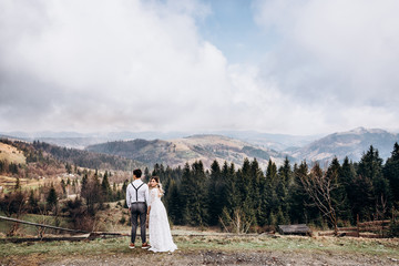 Rear of the wedding couple standing at the very picturesque place in the mountains. Bride looking at the camera over her shoulder. Back view.