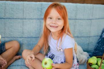 Fototapeta premium Beautiful caucasian girl of five years old eats green apples in the garden, looks into the camera and smiles. Happy childhood