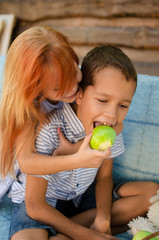 Fototapeta premium Happy boy and girl on a swing in the garden. Sister feeds her brother green apples. Laughing children, happy childhood