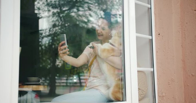 Student pretty girl is taking selfie with pet dog shiba inu breed using smartphone camera sitting in cafe on window sill, animal is licking woman's face.