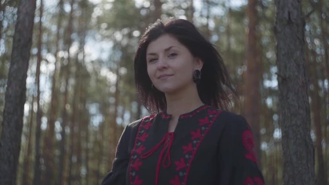 Cute caucasian young woman in black and red dress standing in the pine forest looking to camera. Connection with nature. Slow motion.