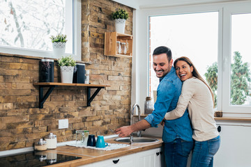 Husband washing dishes and wife hugging him in the modern cozy kitchen.