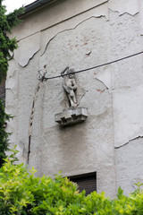 Little boy bearing treasure sculpture, standing on a grungy old gray wall, hidden under an electrical cable, surrounded by leafy green garden 