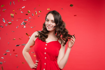 Studio portrait of beautiful young brunette lady with wavy hair in summer red dress with buttons smiling at camera. She is sprinkled or showered with sparkling confetti on red background. Isolate.