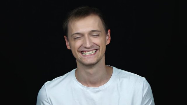 Handsome young man in t-shirt smiling while standing against black background. Male laughs from ear to ear, giggles on the funny stories. False or fake smile