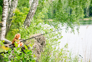 Naklejka na ściany i meble Teddy bear fisherman. Brown teddy bear sits by the lake with a fishing rod and catches fish.