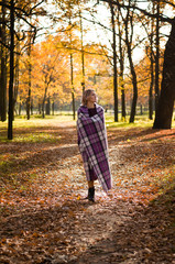 Obraz premium Young beautiful blonde woman wrapped in checkered blanket or plaid walks outdoors through the autumn park at sunset. Comfort and cosiness concept.