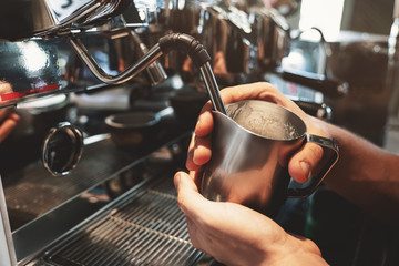 barista man steaming milk in frothing pitcher using professional coffee machine cappuccinator in...