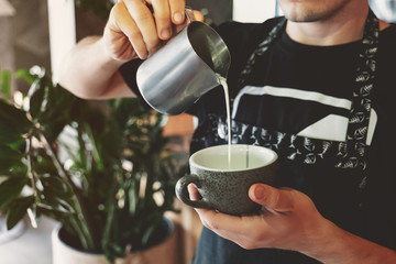 barista man pouring whipped milk from frothing pitcher in cup with coffee in cafe