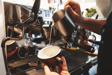 barista man pouring whipped milk from frothing pitcher in cup with coffee standing near...