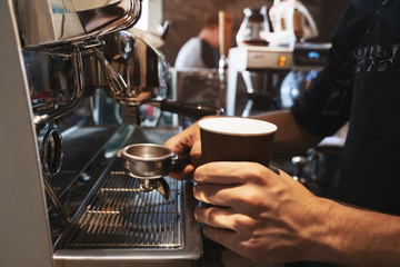 barista man holding hot coffee drink in paper cup in one hand and coffee holder in another standing near professional coffee machine in close up
