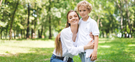 Fototapeta premium panoramic shot of beautiful smiling mom hugging son in park during daytime