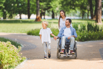 mother rolling wheelchair with disabled father in park near son