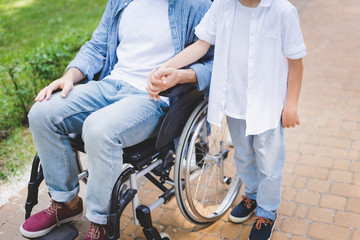 cropped view of son hloding hands with disabled father in wheelchair