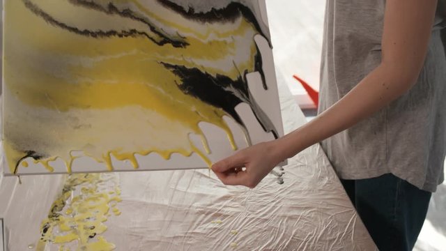 Handheld camera shot of professional female painter tilting canvas with acrylic paints flowing over edges while making fluid painting in art studio