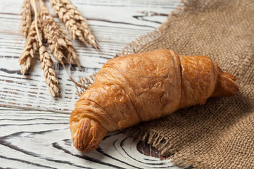One whole baked croissant near bunch of wheat on brown sackcloth and old white rustic wooden table