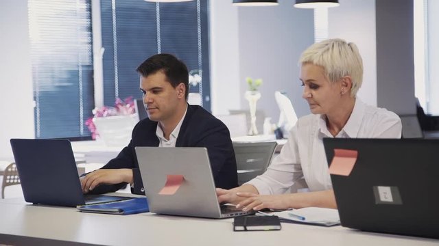 Two mature, qualified and professional colleagues sitting indoor modern office with loft interior work place. Woman and man spending working day using laptop computer and testing program